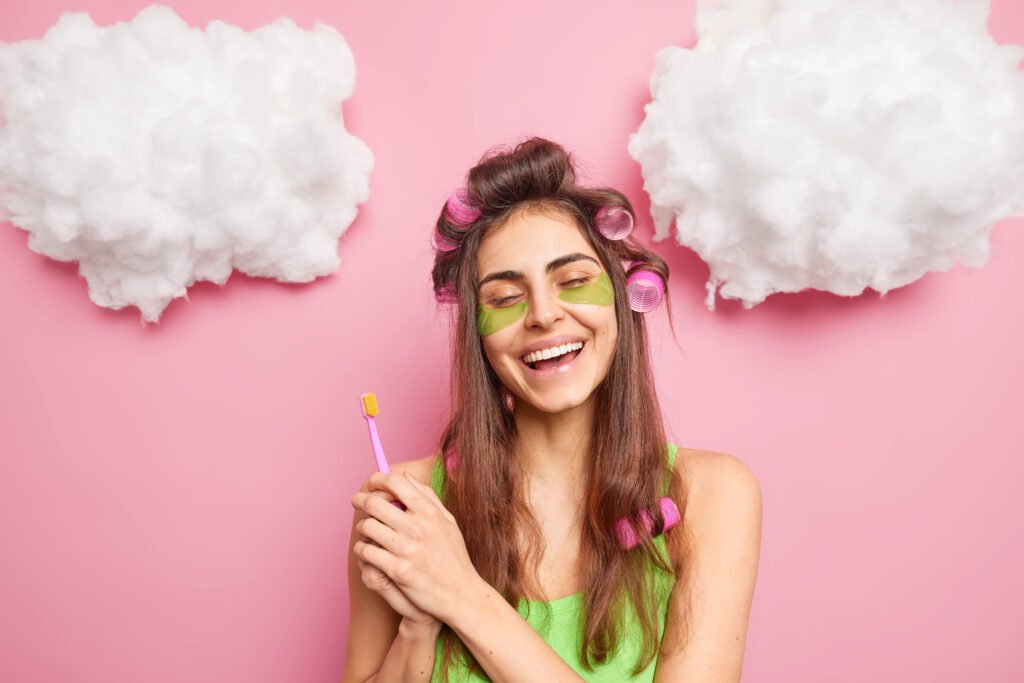 Positive carefree girl makes perfect hairstyle holds toothbrush going to clean teeth enjoys morning routines cares abot skin uses collagen pads smiles toothily isolated over pink background clouds up